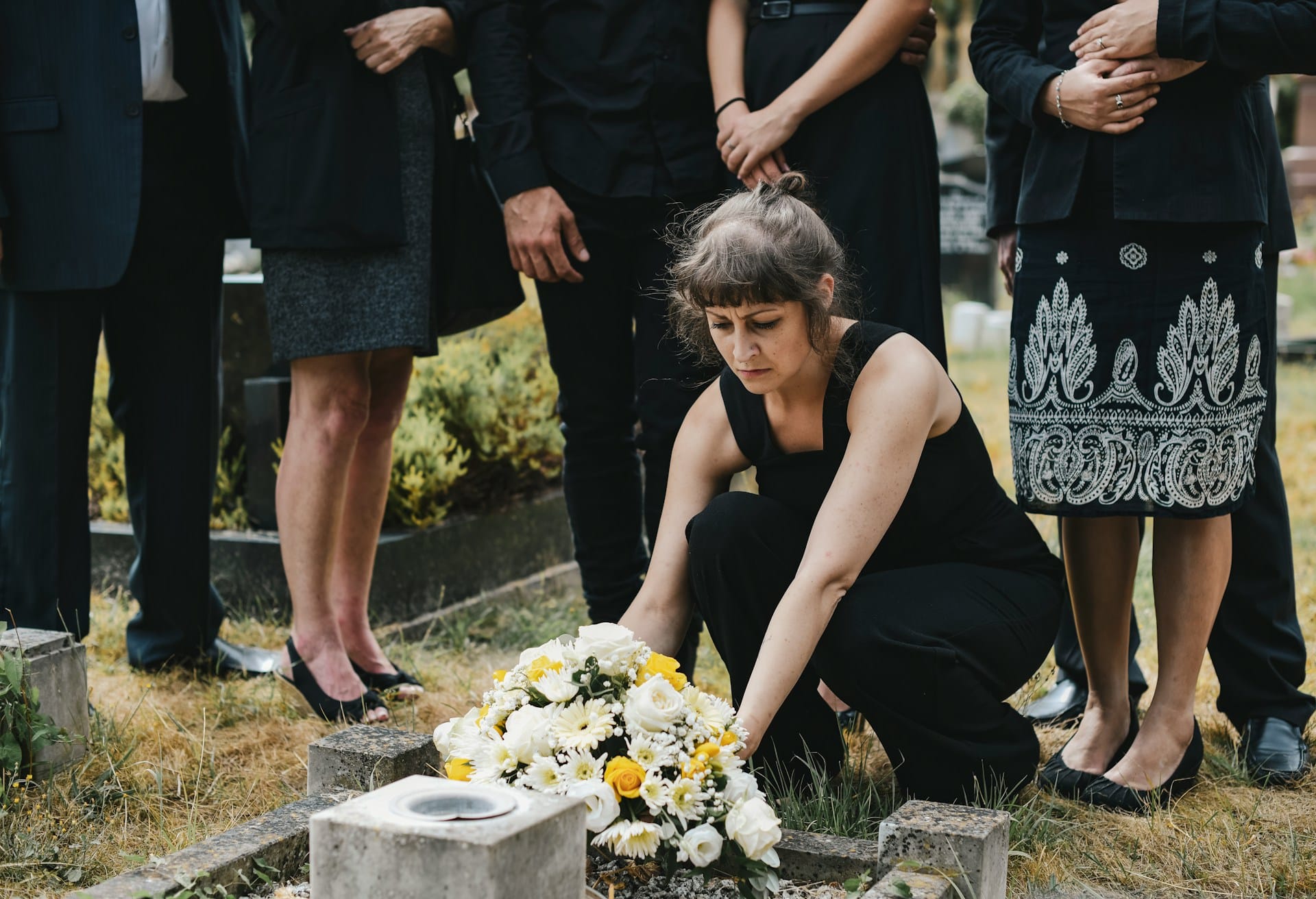 A mourning woman kneels and places flowers on a grave while her family stands behind her with only their lower bodies visible, representing dependant relief claims.