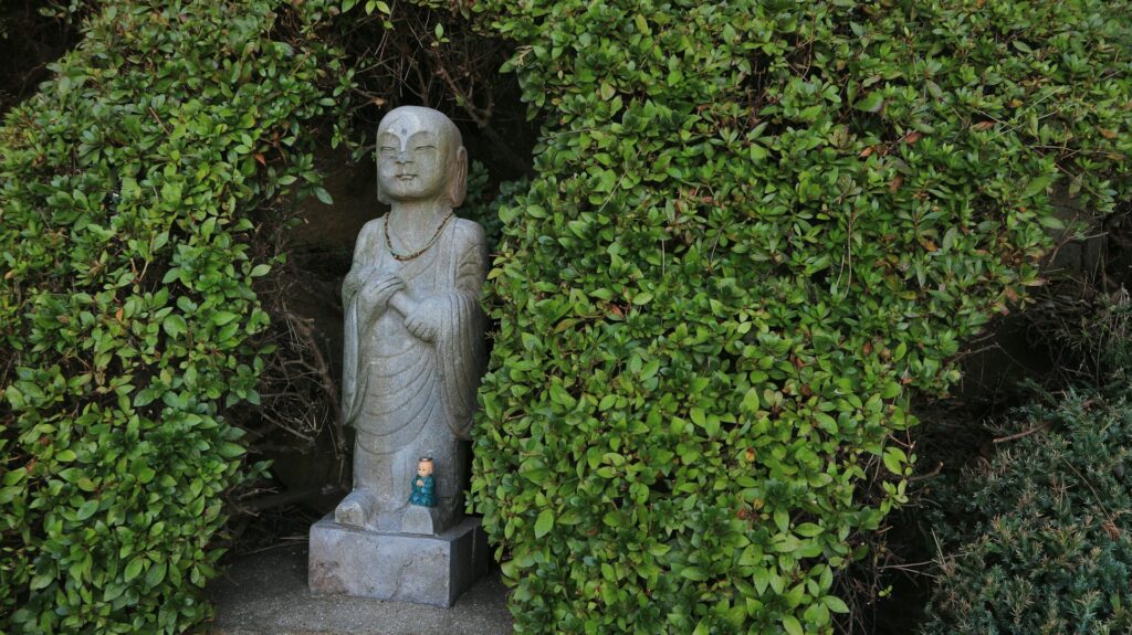 A small, serene stone Buddha statue stands nestled within a dense, bright green hedge. The statue is wearing a simple robe and a small beaded necklace, and it has a calm expression. At the base of the statue is a smaller, teal-colored figurine. The setting evokes a sense of peace and contemplation, contrasting the emotional weight of a final divorce order.
