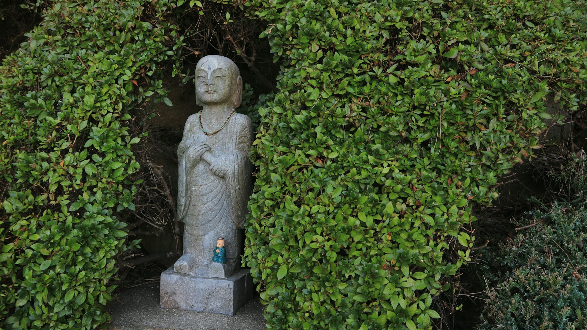 A small, serene stone Buddha statue stands nestled within a dense, bright green hedge. The statue is wearing a simple robe and a small beaded necklace, and it has a calm expression. At the base of the statue is a smaller, teal-colored figurine. The setting evokes a sense of peace and contemplation, contrasting the emotional weight of a final divorce order.
