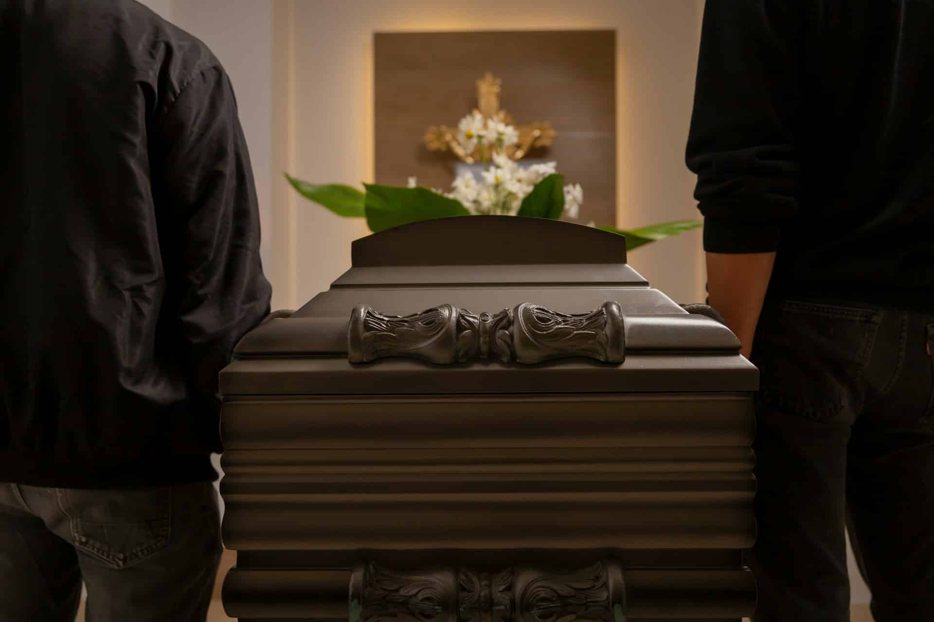 Two men, possibly pallbearers, dressed in dark clothing and visible only from the shoulders to thighs, flank an ornately carved wood casket, representing international estate disputes.