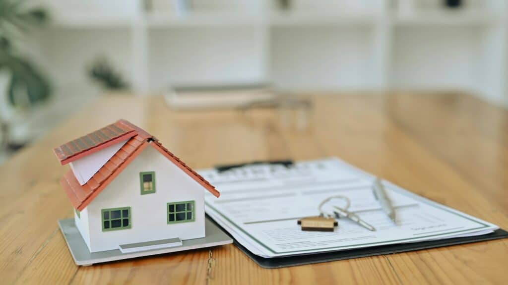 A miniature model of a house is placed next to a legal document, a pen, and a set of keys on a wooden desk. The image illustrates the process of purchasing pre-sale properties and relates to a developer's requirements to disclose crucial information, such as building permits and financing commitments, as well as outlining buyers' rights to terminate agreements.