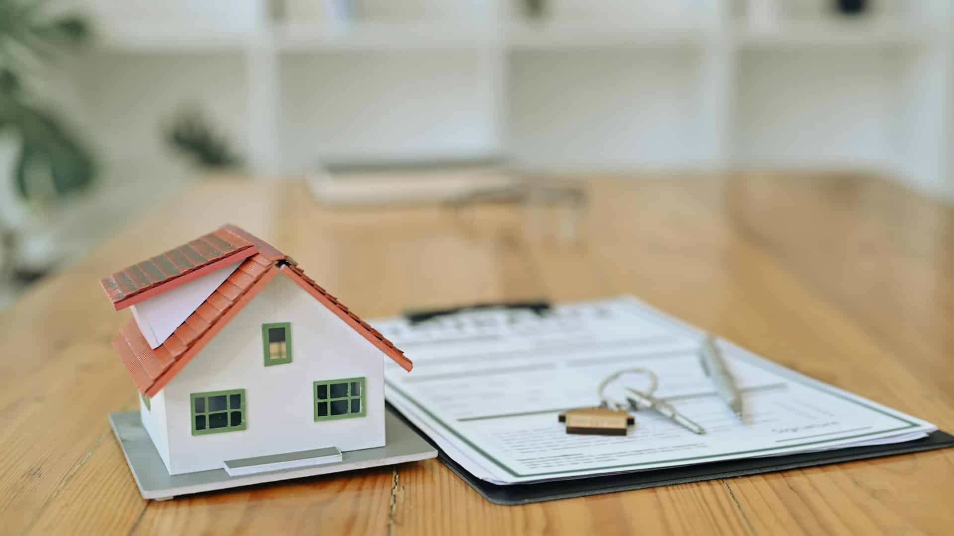 A miniature model of a house is placed next to a legal document, a pen, and a set of keys on a wooden desk. The image illustrates the process of purchasing pre-sale properties and relates to a developer's requirements to disclose crucial information, such as building permits and financing commitments, as well as outlining buyers' rights to terminate agreements.
