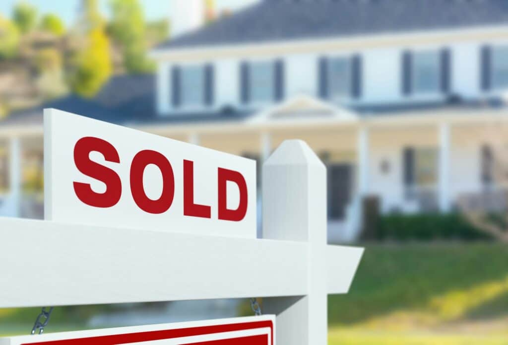 A sold sign in front of a large, white family home blurred in the background, with a meticulous green lawn and tall trees growing to the side of the house, representing BC Flipping Tax.