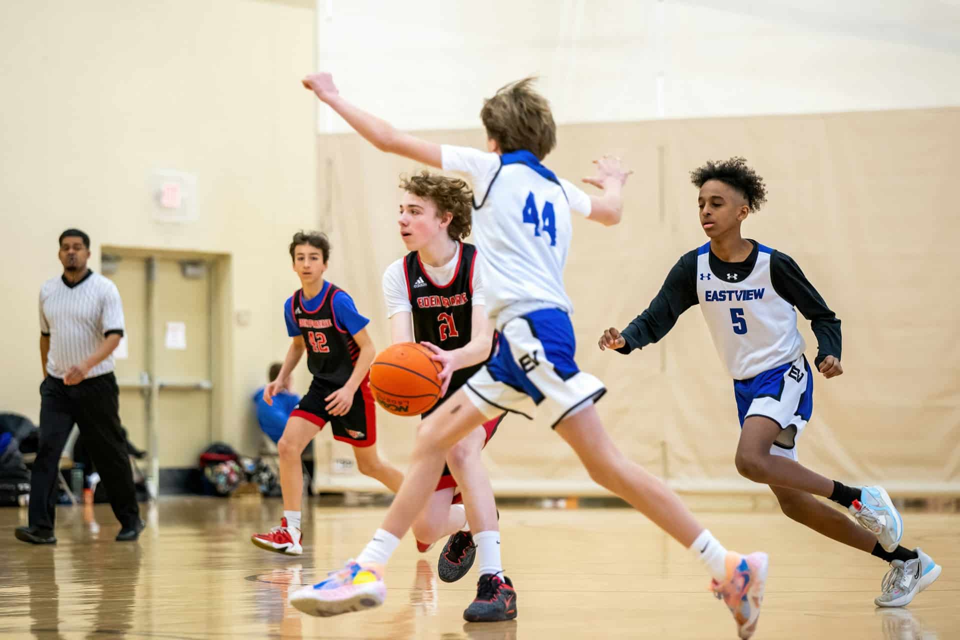 Four teenage boys are playing basketball in a gymnasium with a referee in the background, representing amateur and youth sports organizations in BC.