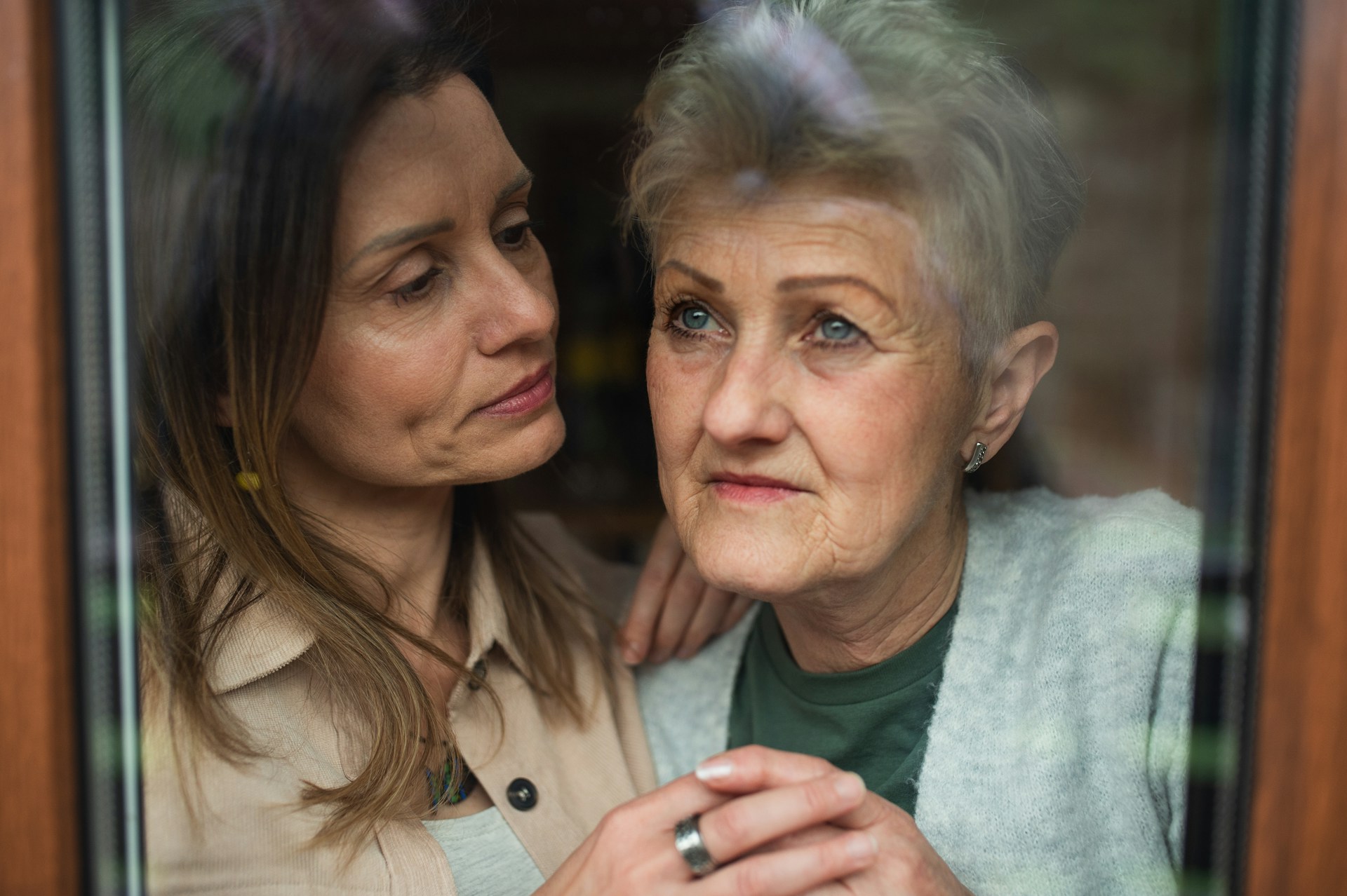 An elderly woman looks out a window, while her daughter stands next to her holding her hand, her expression sad, representing emergency asset protection and committeeship.