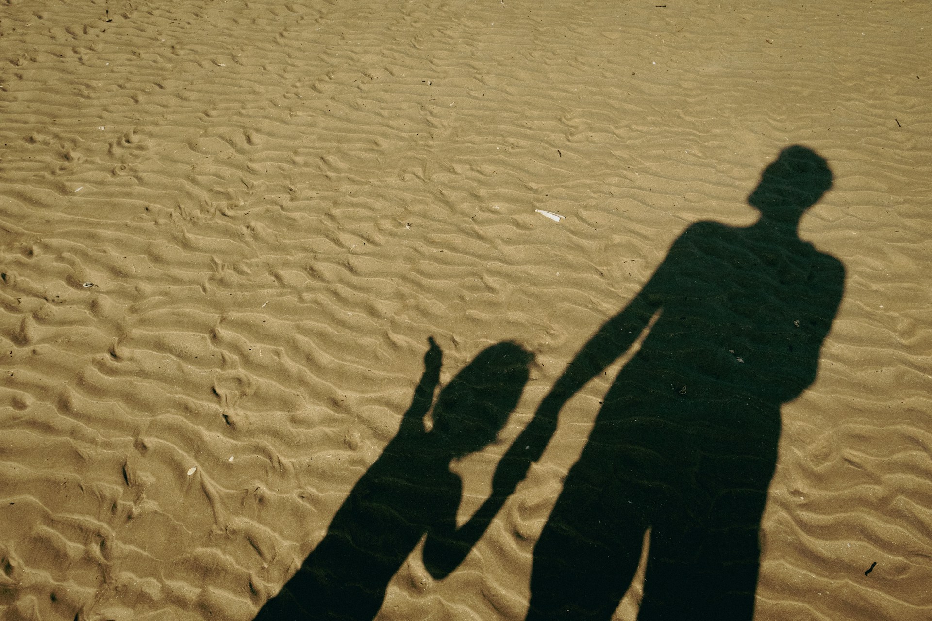 Shadows of a parent holding their child's hand stretched out over a rippled sandy beach, representing parental alienation.