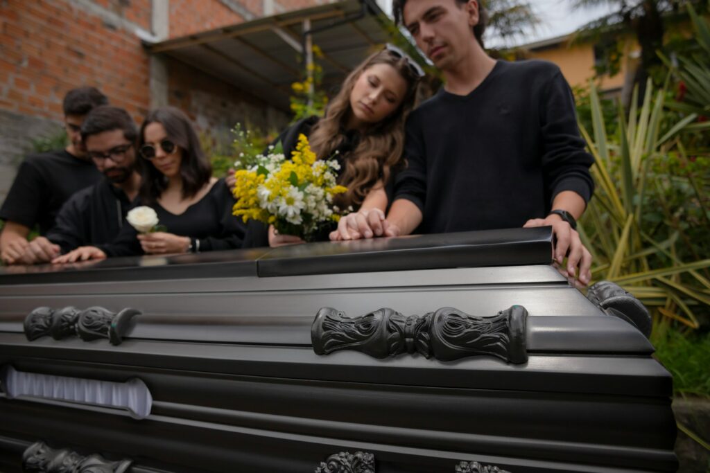 Mourning adult children, three men and two women, rest their hands on a dark wood casket with one woman holding a bouquet of yellow an white flowers and the other holding a white rose, representing disinheriting a child.