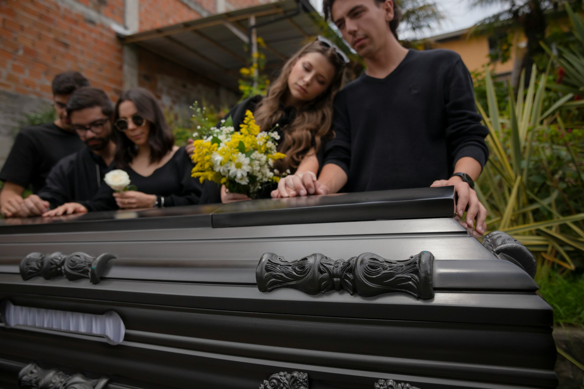 Mourning adult children, three men and two women, rest their hands on a dark wood casket with one woman holding a bouquet of yellow an white flowers and the other holding a white rose, representing disinheriting a child.