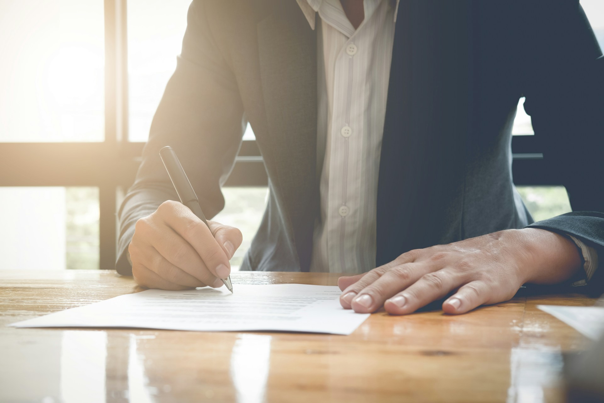 A man, shown from the shoulders down, signs paperwork on a wooden table, while the sun casts a golden glow through the windows behind him, representing pre-Family Law Act marriage agreements in BC.