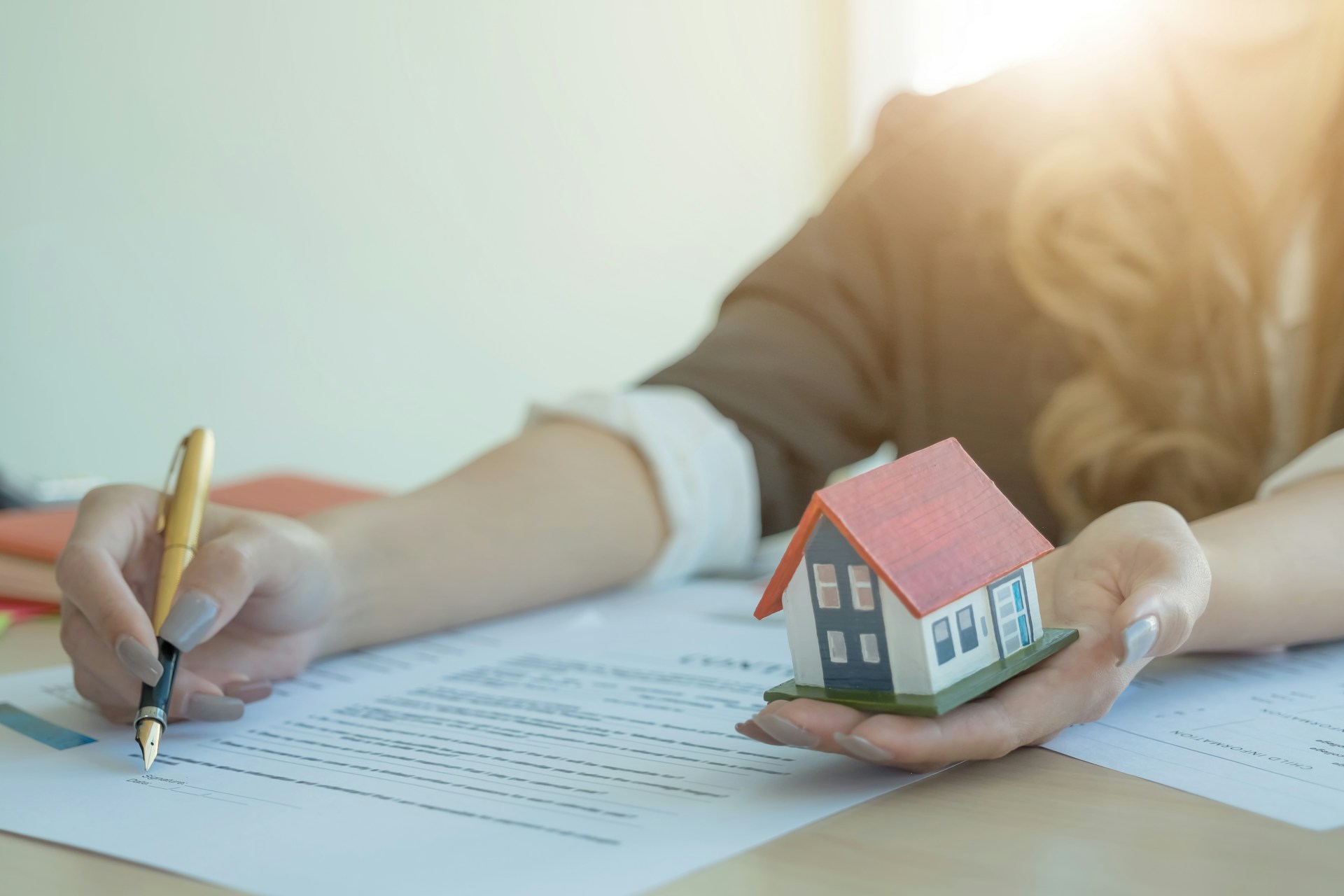 A woman is signing paperwork while holding onto a small model home that represents real estate fraud in Vancouver.