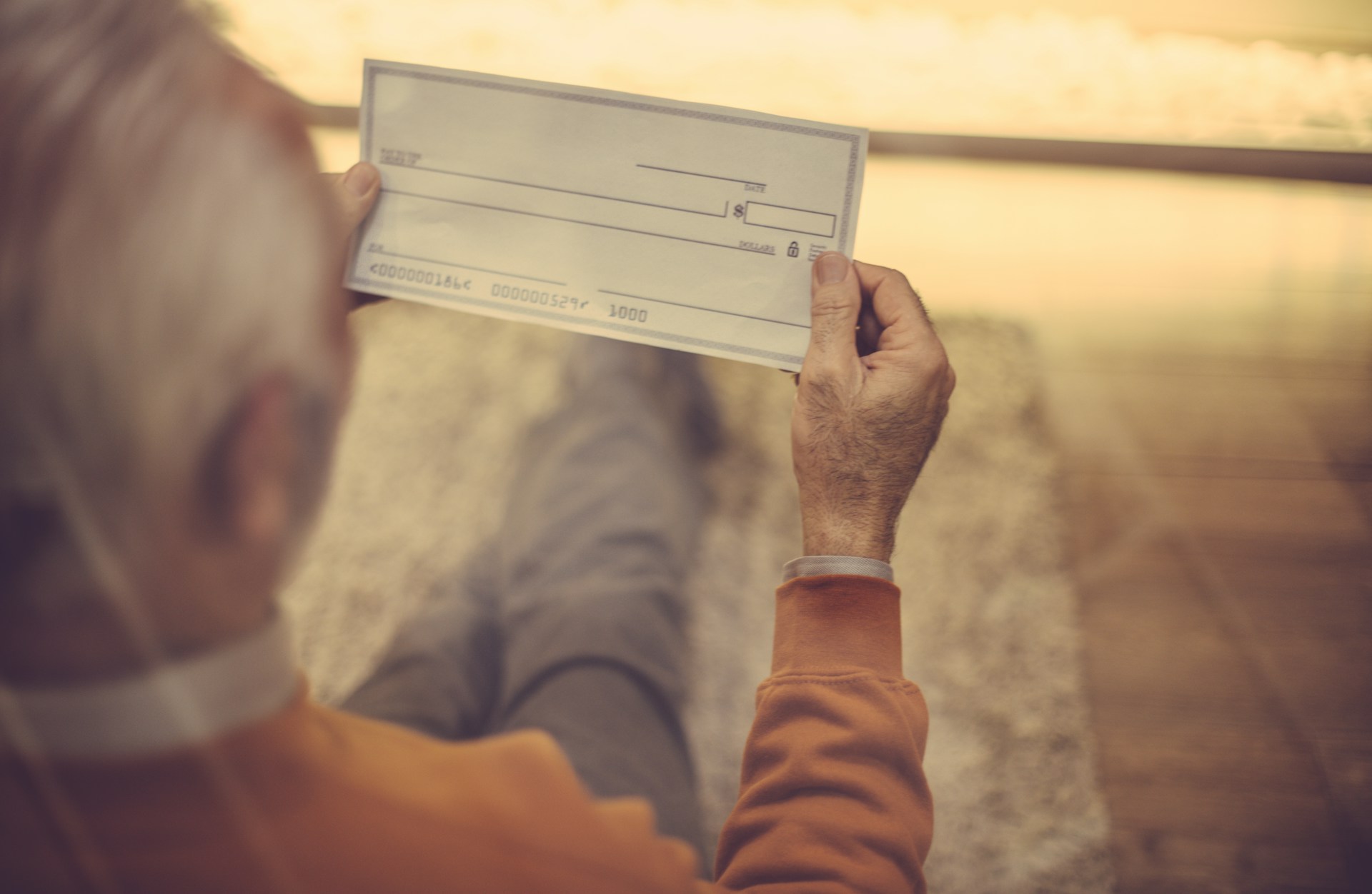 An elderly man, sitting with his legs crossed at the ankles, looks down at the blank cheque he holds pinched between the fingers of both hands, representing late-life gifts and elder abuse.