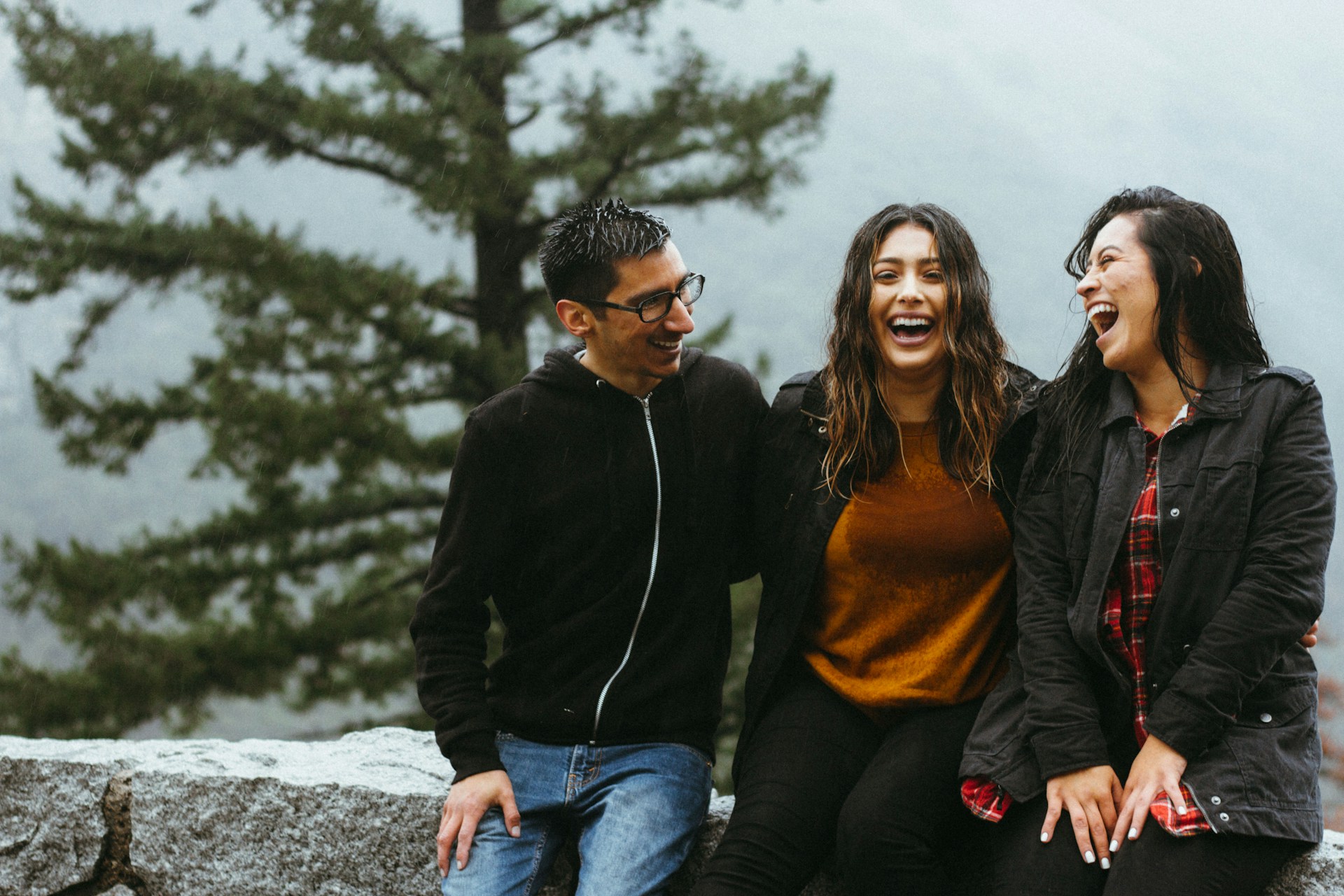 A man and two women sit together on a stone wall, laughing on a rainy day with a large pine tree in the background, representing polyamorous relationships in BC.