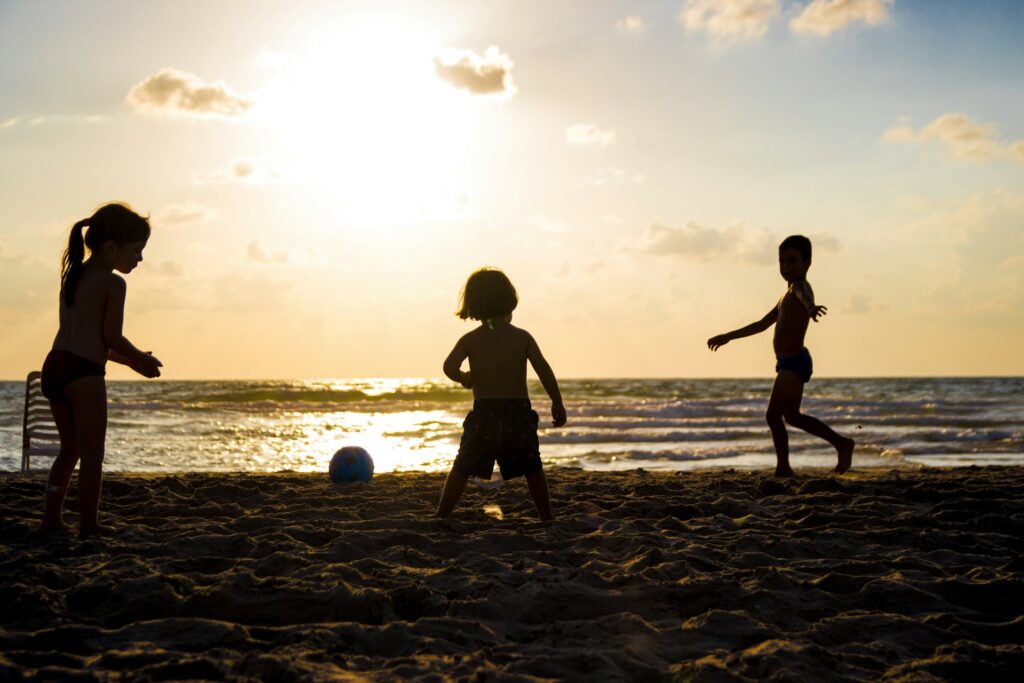 Three young children are silhouetted against the setting sun while they play with a ball in the sand at the beach, representing summer parenting schedules in BC.