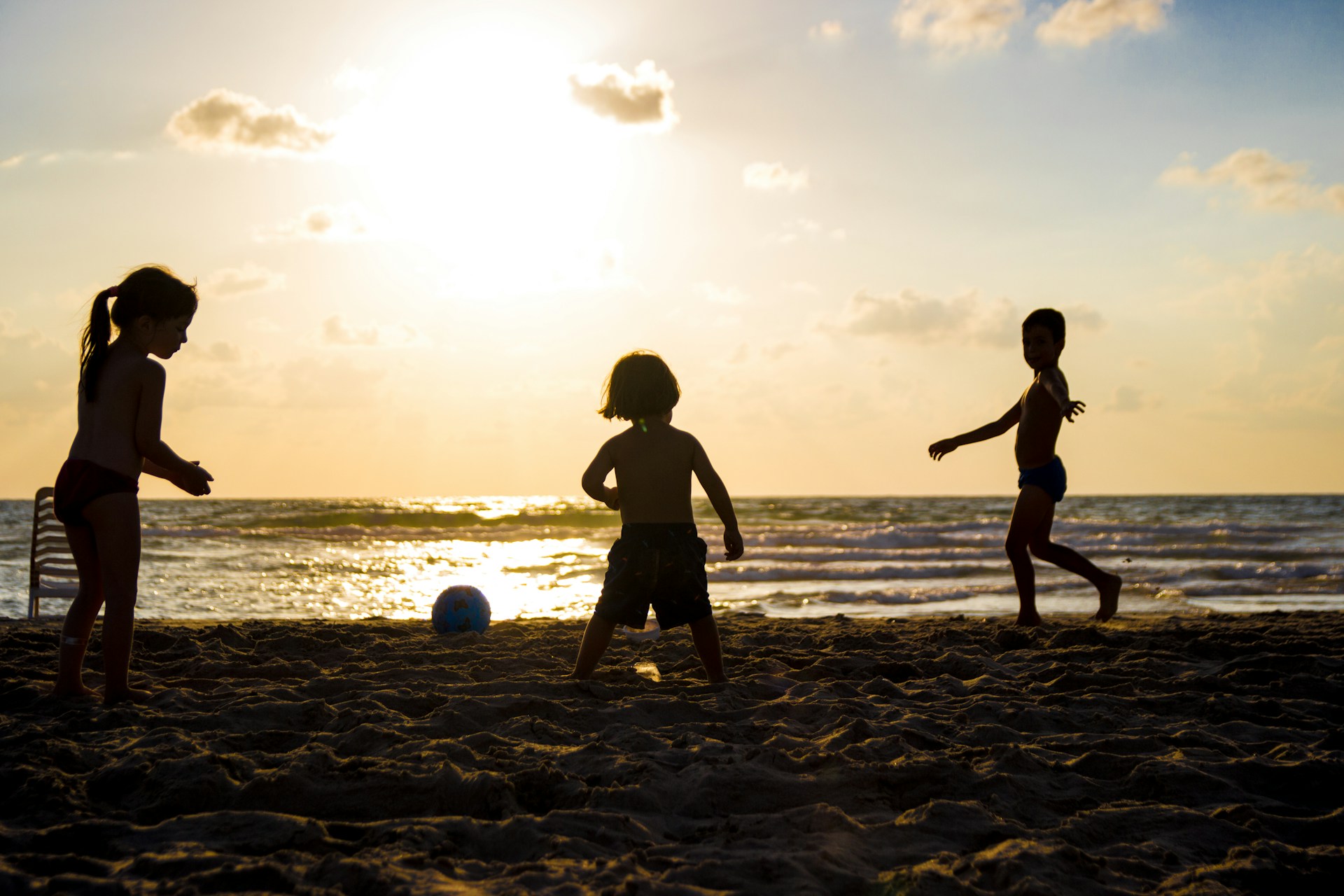Three young children are silhouetted against the setting sun while they play with a ball in the sand at the beach, representing summer parenting schedules in BC.