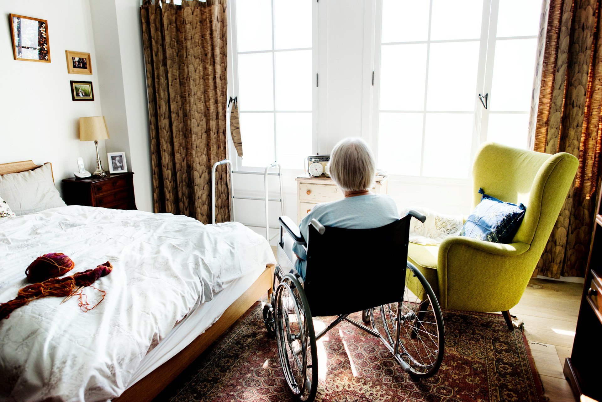 An elderly woman in a wheelchair looks out her bedroom windows positioned between a chair and her bed with her knitting on the corner, representing undue influence will challenges.