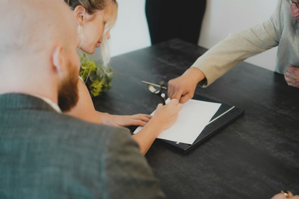 A man and a woman sign a prenuptial agreement presented to them by a person sitting opposite them at a desk, representing prenups in British Columbia.